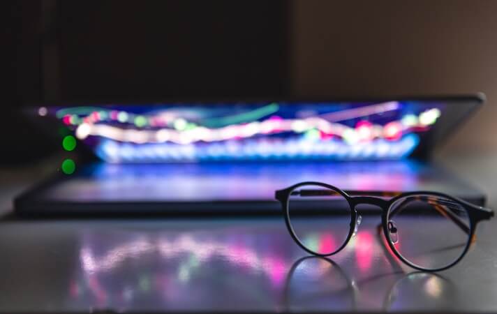 Glasses sat on a desk in front of a partially closed laptop showing digital investment information in bright colours. 